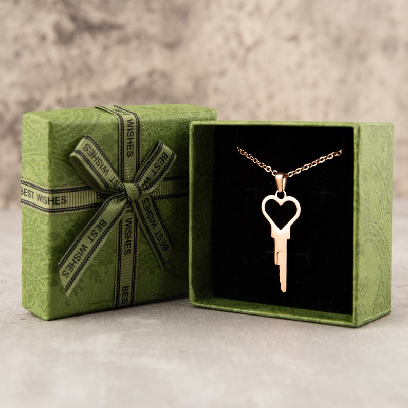 Puede incluir: Un collar de llave de coraz&oacute;n de oro en una caja de regalo verde con interior de terciopelo negro. La caja est&aacute; decorada con una cinta verde y las palabras "Mejores deseos" impresas en ella.