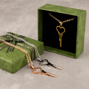 Puede incluir: Tres collares de oro con colgantes de llave en forma de coraz&oacute;n en una caja de regalo verde. La caja tiene las palabras "Mejores deseos" impresas.