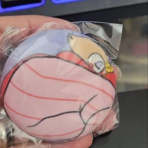May include: A round, pink plushie with a cartoon character design, wrapped in clear plastic. The plushie features a sleeping figure with red and black lines, and a yellow and orange character.