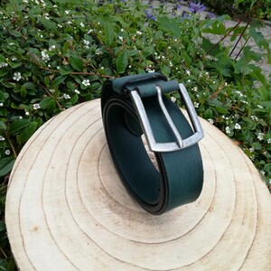 May include: A dark green leather belt with a silver buckle. The belt is rolled up and sitting on a wooden surface.