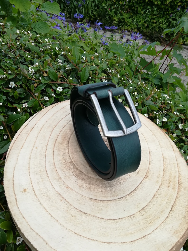 May include: A dark green leather belt with a silver buckle. The belt is rolled up and sitting on a wooden surface.
