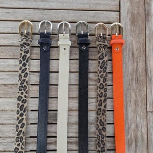 Könnte beinhalten: Sechs Gürtel in verschiedenen Farben und Mustern werden auf einer Holzoberfläche gezeigt. Die Gürtel umfassen Leopardenmuster, Marineblau, Creme, Schwarz, Leopardenmuster und Orange. Jeder Gürtel hat eine Metallschnalle.