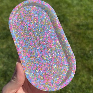 May include: An oval-shaped resin tray filled with colorful glitter. The tray is clear with a multitude of pink, blue, and yellow glitter pieces. The tray is held in a person's hand against a backdrop of green grass.