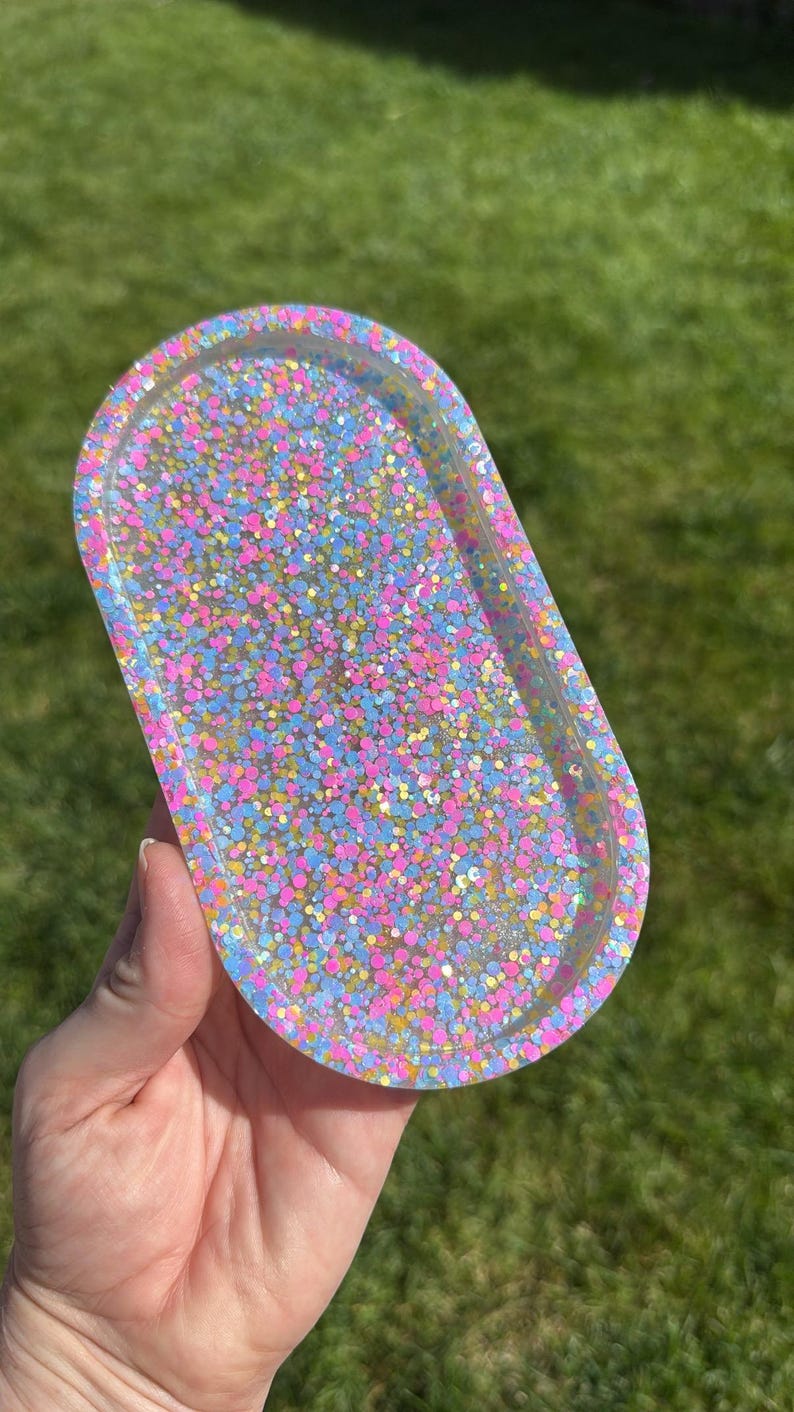 May include: An oval-shaped resin tray filled with colorful glitter. The tray is clear with a multitude of pink, blue, and yellow glitter pieces. The tray is held in a person's hand against a backdrop of green grass.