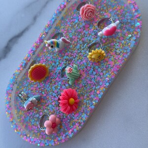 May include: A collection of colorful rings with various designs, including a unicorn, flowers, and a sun, displayed on a glittery, oval-shaped tray. The rings are made of plastic and have silver-colored bands.