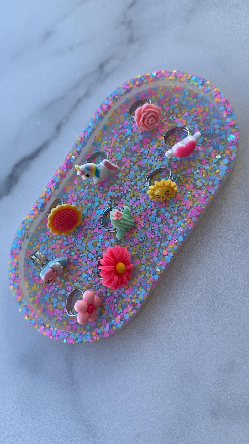 May include: A collection of colorful rings with various designs, including a unicorn, flowers, and a sun, displayed on a glittery, oval-shaped tray. The rings are made of plastic and have silver-colored bands.