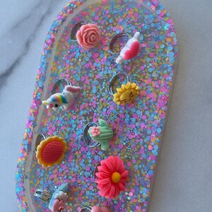 May include: A collection of colorful rings with various designs, including flowers, a unicorn, and a sun, set on a glittery, oval-shaped tray. The rings are made of resin and have silver-colored bands.