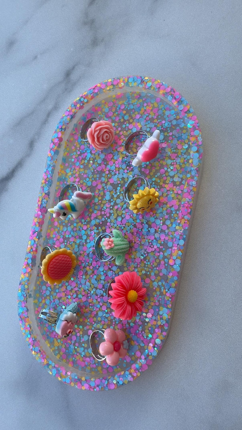 May include: A collection of colorful rings with various designs, including flowers, a unicorn, and a sun, set on a glittery, oval-shaped tray. The rings are made of resin and have silver-colored bands.