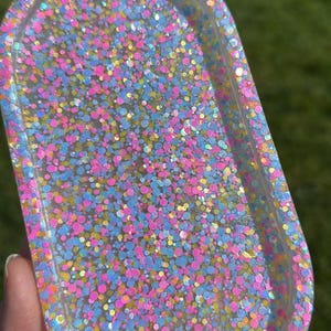 May include: An oval-shaped resin tray filled with colorful glitter. The glitter is a mix of pink, blue, yellow, and iridescent specks. The tray is held in a person's hand, with a green background visible.