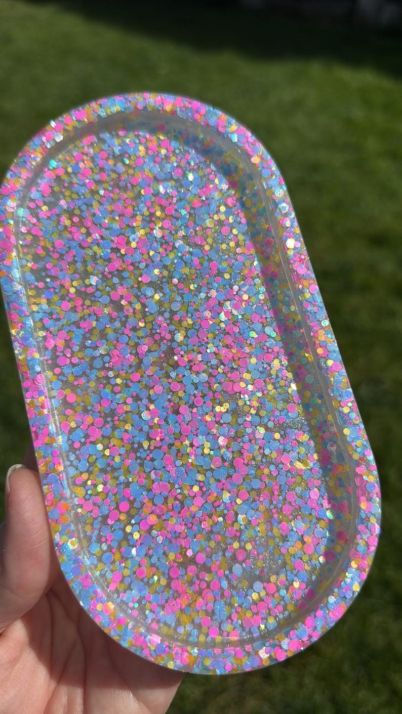 May include: An oval-shaped resin tray filled with colorful glitter. The glitter is a mix of pink, blue, yellow, and iridescent specks. The tray is held in a person's hand, with a green background visible.