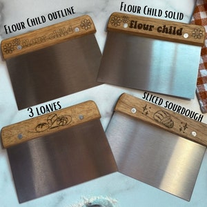 May include: Four different wooden handled dough cutters with various designs. The designs include "Flour Child" with a flower outline, "Flour Child" with a solid flower design, "3 Loaves" with a bread design, and "Sliced Sourdough" with a bread design. A small metal sifter is also in the image.