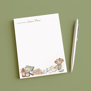 May include: White notepad with a watercolor illustration of baking tools and ingredients. The notepad has the text "A Note From Jessica Marie" in brown script. A silver pen rests beside the notepad on a green surface.