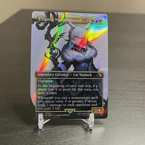 May include: A holographic trading card featuring Y'shtola, Night's Blessed, a Cat Warlock. The card has a rainbow sheen and includes text describing its abilities. The card is displayed on a clear plastic stand.