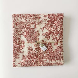 Peut inclure: Un tissu rectangulaire plié avec un motif floral rouge. Le tissu est noué avec de la ficelle et une petite étiquette. Le motif toile de Jouy présente des fleurs, du feuillage et des éléments architecturaux. Le tissu est de couleur crème.