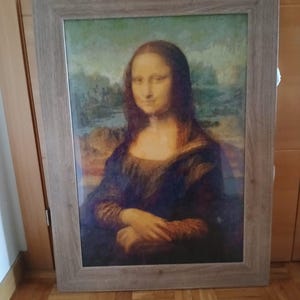 Puede incluir: Una impresión enmarcada de la pintura de la Mona Lisa. La obra presenta un retrato de una mujer con una sonrisa sutil, vestida con un vestido oscuro. El marco es de color marrón claro y el fondo muestra un paisaje.