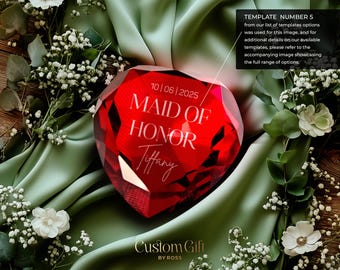 May include: A red crystal heart-shaped award with the text "10/06/2025 Maid of Honor Tiffany" engraved on it. The award is sitting on a green fabric surface with white flowers.