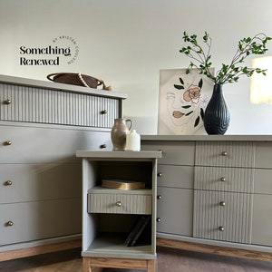 May include: A set of three gray dressers with fluted fronts and brass knobs. The dressers are arranged in a row, with a smaller dresser in the middle and two larger dressers on either side. The dressers are all on a wooden floor. The smaller dresser has a shelf and a drawer. The larger dressers have three drawers each. The dressers are decorated with a vase of flowers and a framed print.