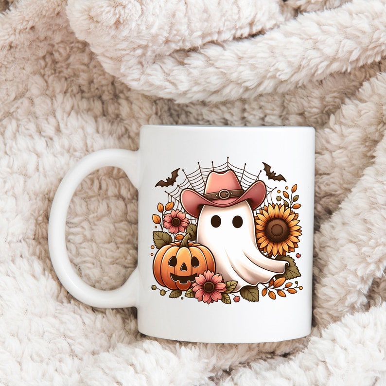 May include: White ceramic mug featuring a colourful illustration of a ghost wearing a pink cowboy hat, a pumpkin, and sunflowers. The ghost has two black eyes and a mouth.