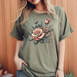 May include: Olive green t-shirt featuring a floral design with roses and leopard print accents. The graphic includes detailed roses in shades of brown and pink, surrounded by green leaves and wheat stalks. The shirt has short sleeves and a crew neck.