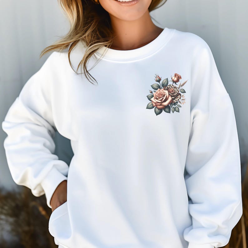 May include: White sweatshirt featuring a floral design on the upper left chest. The floral arrangement includes roses in shades of peach and brown, with green leaves and a touch of leopard print. The sweatshirt has a crew neck and long sleeves.