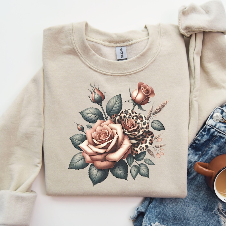 May include: A beige sweatshirt featuring a floral design with roses in shades of peach and a leopard print accent. The design includes green leaves and buds. The sweatshirt is made of a soft material.