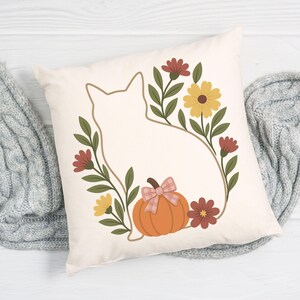 May include: Cream-colored square throw pillow featuring a cat silhouette surrounded by a floral wreath. The wreath includes yellow and maroon flowers, green leaves, and an orange pumpkin with a pink plaid bow. The pillow is a fall-themed decorative item.