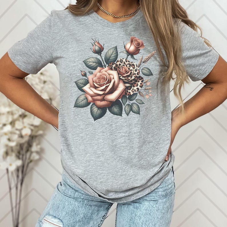 May include: A gray t-shirt featuring a floral design. The design includes roses in shades of pink and brown, with green leaves and a leopard print accent. The shirt has a round neckline and short sleeves.