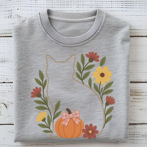 May include: A light gray t-shirt featuring a cat silhouette surrounded by a floral wreath. The wreath includes red, yellow, and brown flowers, green leaves, and an orange pumpkin with a pink bow. The shirt is folded on a white wooden surface.