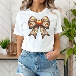 May include: White t-shirt featuring a large, colourful bow with a pumpkin in the centre. The bow is made of ribbon with a floral pattern and orange and green accents.