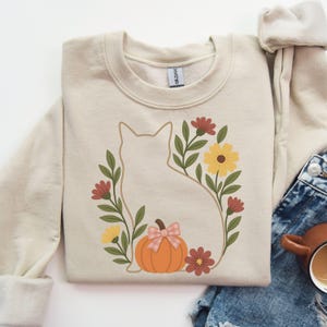 May include: Beige crewneck sweatshirt featuring a cat silhouette surrounded by a floral wreath. The wreath includes red, yellow, and green flowers, and a pumpkin with a pink bow. A cozy fall-themed design.