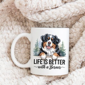 May include: White ceramic coffee mug with a colorful illustration of a Bernese Mountain Dog peeking over a wooden sign. The text "Life is better with a Berner" is printed on the sign.