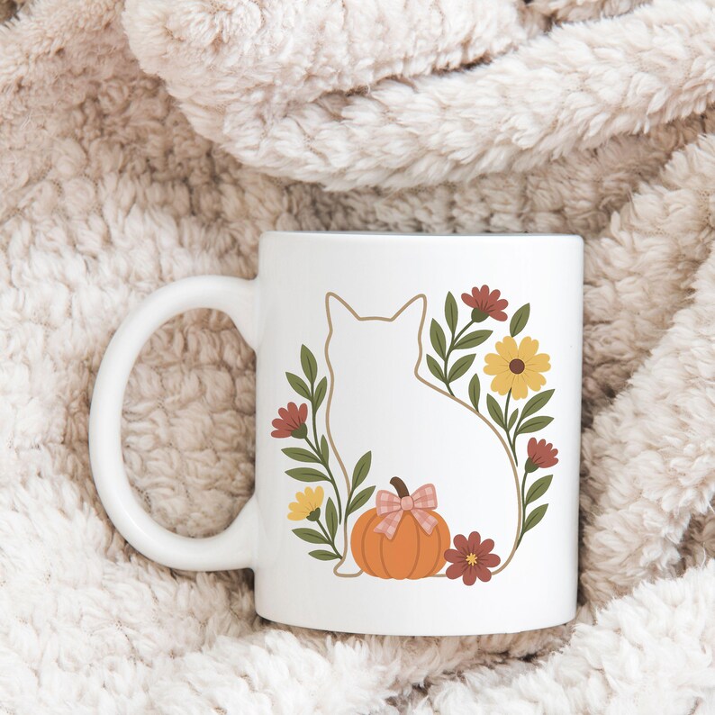 May include: White ceramic mug featuring a cat silhouette surrounded by a floral wreath with a pumpkin and bow. The design includes orange, yellow, brown, and green elements. The mug has a curved handle and is set against a soft, textured background.