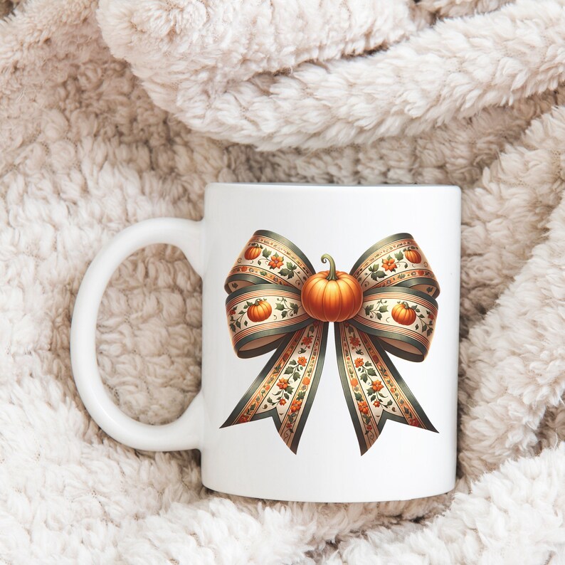 May include: White ceramic mug featuring a green and orange bow with a pumpkin at its centre. The bow is adorned with small pumpkins and floral patterns.