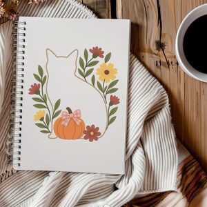 May include: A spiral-bound notebook featuring a cat silhouette surrounded by a floral wreath with a pumpkin and bow. The design includes yellow and red flowers, green leaves, and an orange pumpkin. The notebook is on a striped blanket.