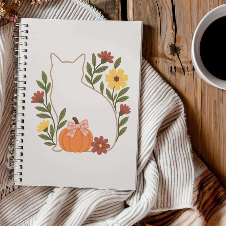 May include: A spiral-bound notebook featuring a cat silhouette surrounded by a floral wreath with a pumpkin and bow. The design includes yellow and red flowers, green leaves, and an orange pumpkin. The notebook is on a striped blanket.