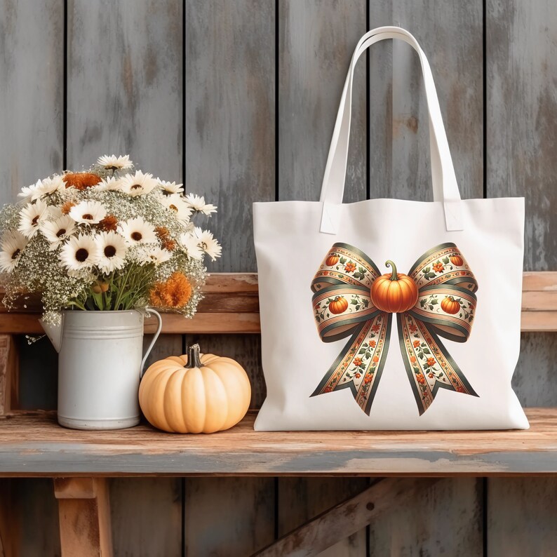 May include: A white tote bag featuring a large, green and orange bow with a pumpkin at its centre. The bow is adorned with small pumpkins and leaves. The bag is resting on a wooden shelf alongside a vase of white flowers and a small pumpkin.