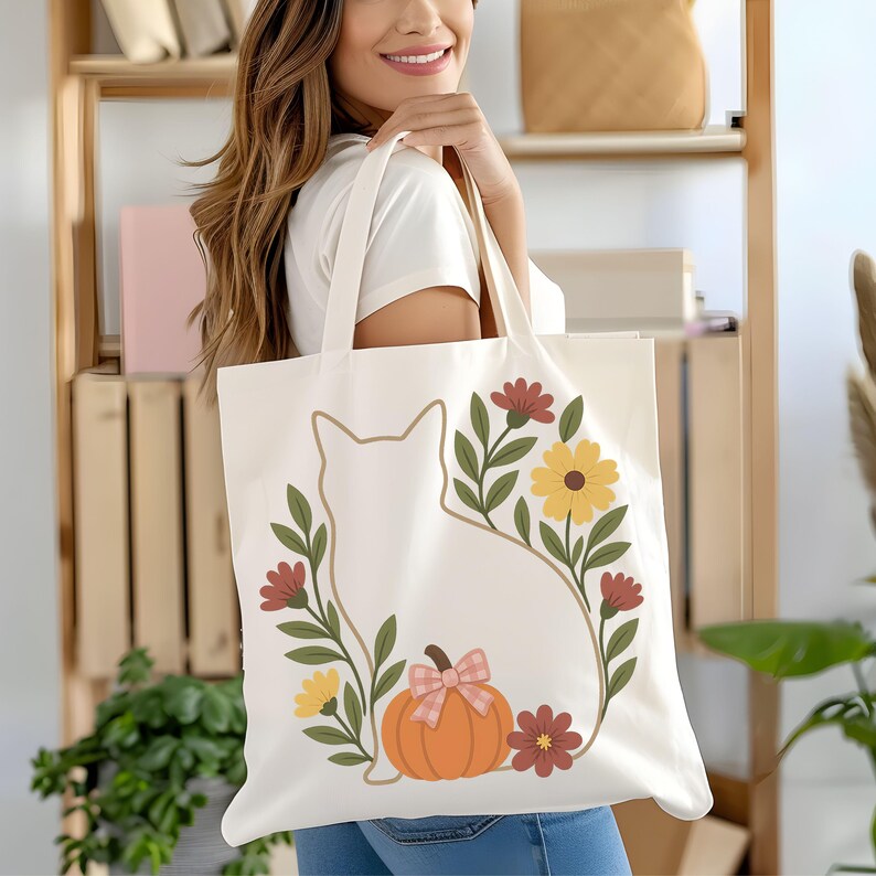 May include: Cream-colored tote bag featuring a cat silhouette surrounded by a floral wreath with a pumpkin and bow. The design includes yellow and maroon flowers, green leaves, and an orange pumpkin with a pink bow. The bag is held by a person.