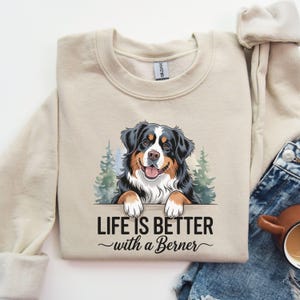 May include: A beige sweatshirt with a graphic of a Bernese Mountain Dog peeking over a sign that reads "Life is better with a Berner".
