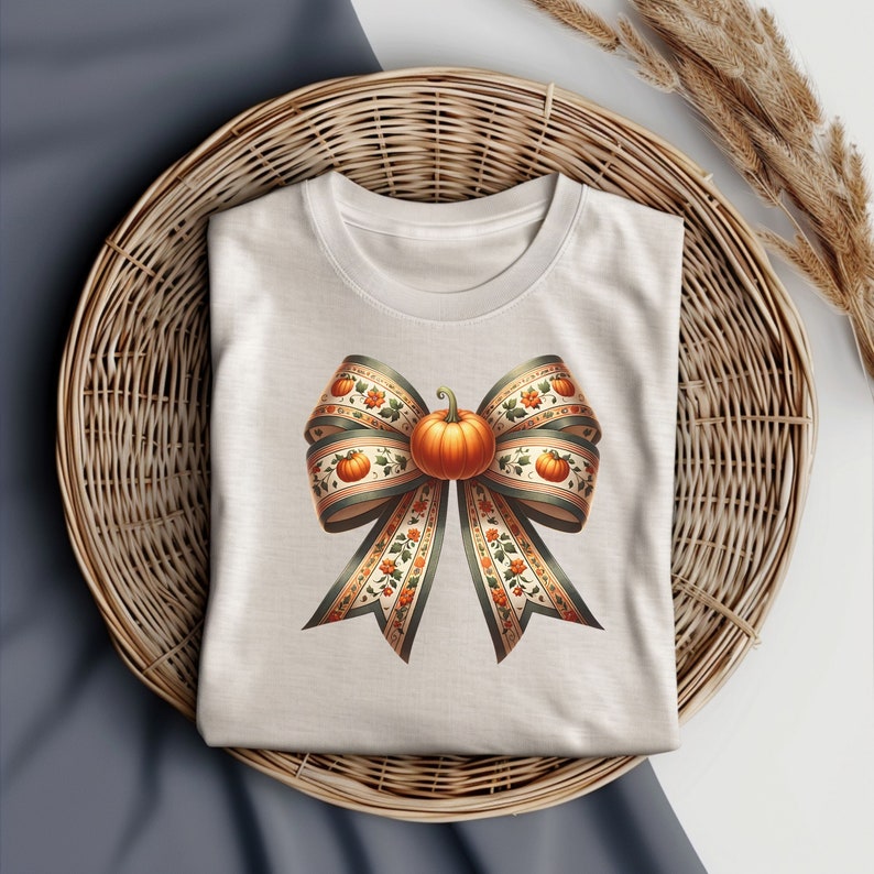 May include: A white t-shirt featuring a large, decorative bow made of fabric with a pumpkin design. The bow is tied in the centre with a small pumpkin in the middle.