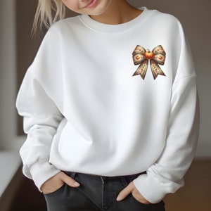 May include: White sweatshirt featuring a large brown and orange bow with a pumpkin in the centre. The bow is made of fabric with a pumpkin pattern.