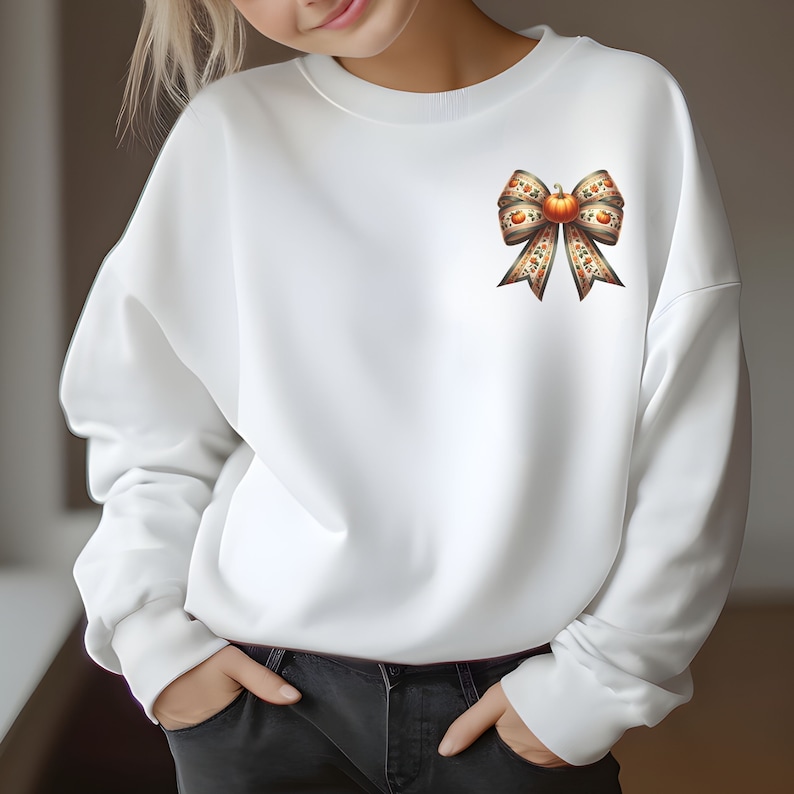 May include: White sweatshirt featuring a large brown and orange bow with a pumpkin in the centre. The bow is made of fabric with a pumpkin pattern.