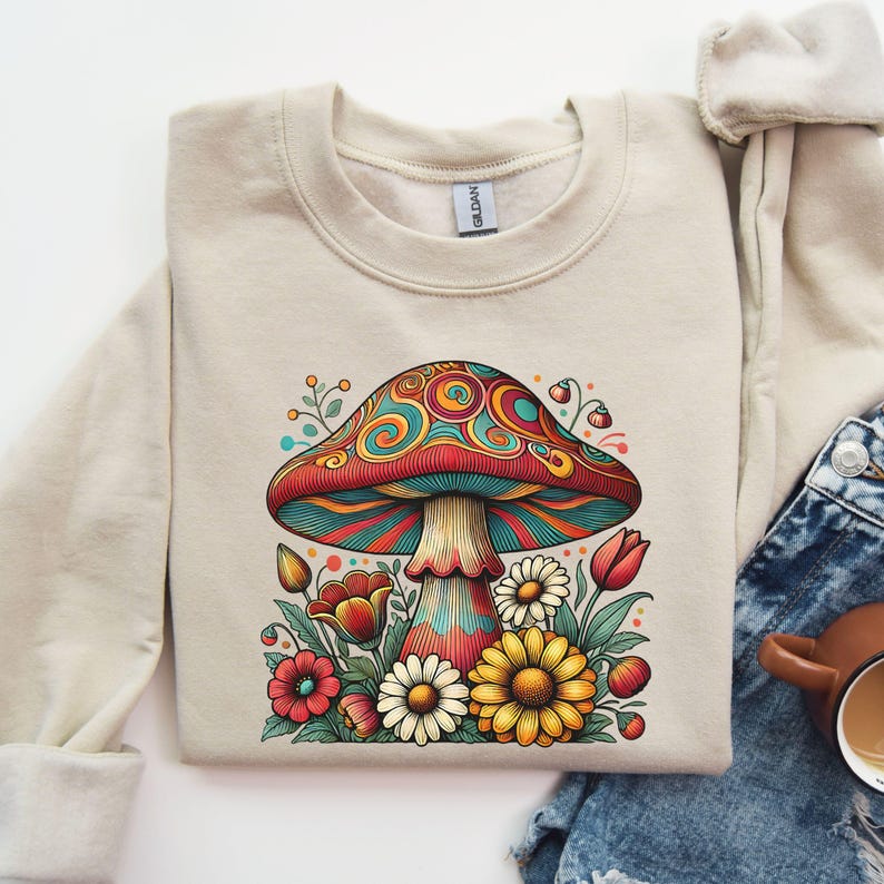 May include: A beige sweatshirt with a colorful psychedelic mushroom graphic surrounded by flowers. The mushroom has a red cap with blue and green swirls and a white stem.