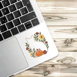May include: A white cat silhouette sticker surrounded by a floral wreath with a pumpkin and bow. The wreath includes red, yellow, and green elements. The sticker is on a laptop, set against a wooden background.