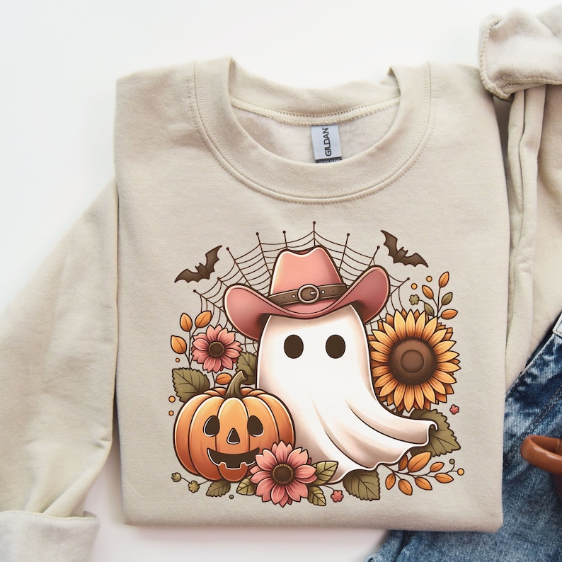 May include: A beige sweatshirt featuring a white ghost wearing a pink cowboy hat and holding a jack-o'-lantern. The ghost is surrounded by sunflowers, leaves, and flowers.
