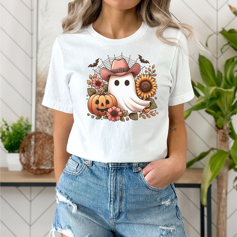May include: White t-shirt featuring a cartoon ghost wearing a pink cowboy hat and holding a jack-o'-lantern. The ghost is surrounded by autumnal flowers and leaves.