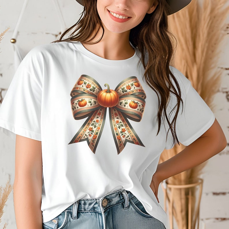 May include: White t-shirt featuring a large, colourful bow made of fabric with a pumpkin in the centre. The bow has a green, orange, and brown pattern.