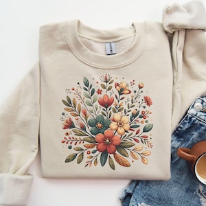 Peut inclure: Un sweat-shirt beige avec un motif floral repr&eacute;sentant un bouquet de fleurs dans des tons de rouge, orange, jaune et vert. Les fleurs sont stylis&eacute;es et ont une esth&eacute;tique vintage.