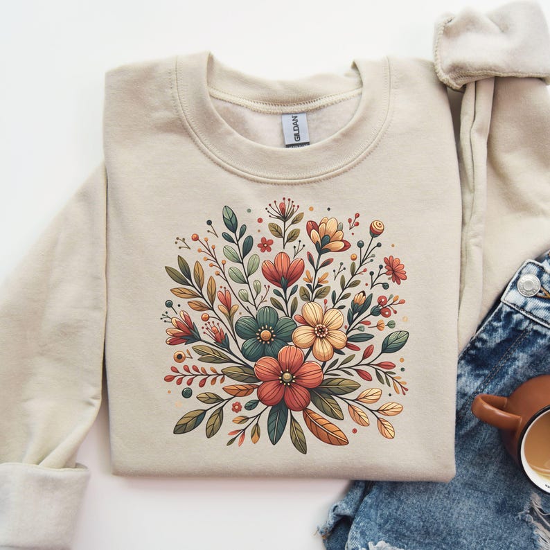 Peut inclure: Un sweat-shirt beige avec un motif floral repr&eacute;sentant un bouquet de fleurs dans des tons de rouge, orange, jaune et vert. Les fleurs sont stylis&eacute;es et ont une esth&eacute;tique vintage.