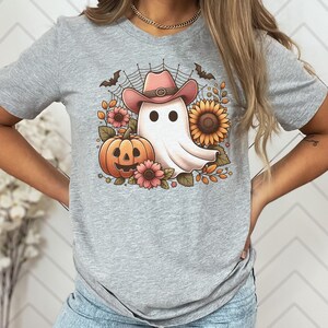May include: A grey t-shirt featuring a white ghost wearing a pink cowboy hat. The ghost is surrounded by autumnal foliage, including a pumpkin, sunflowers, and leaves.