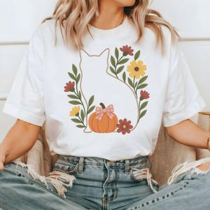 May include: White t-shirt featuring a cat silhouette surrounded by a floral wreath with a pumpkin and bow. The design includes yellow, burgundy, and green elements, creating a fall-themed graphic.